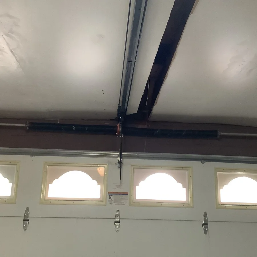 A torsion shaft running the full width of a garage door header above a row of arched window lites, the opener rail crossing beneath a timber ceiling joist.