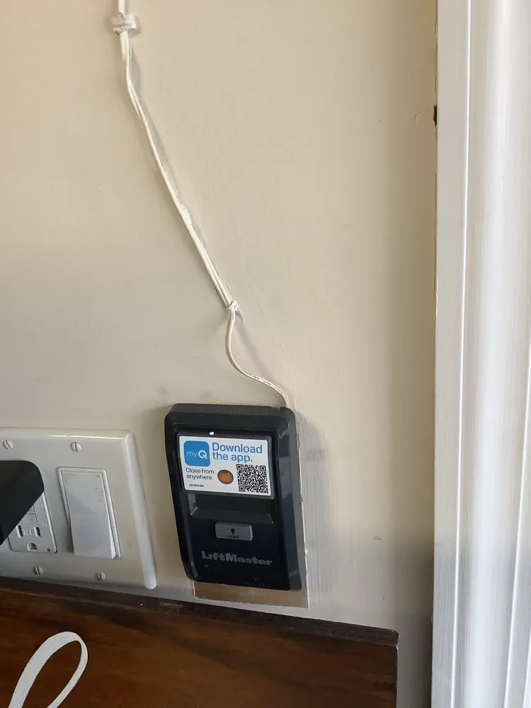 A LiftMaster wall control button mounted beside the light switches on a garage wall, its low-voltage bell wire stapled up towards the ceiling.