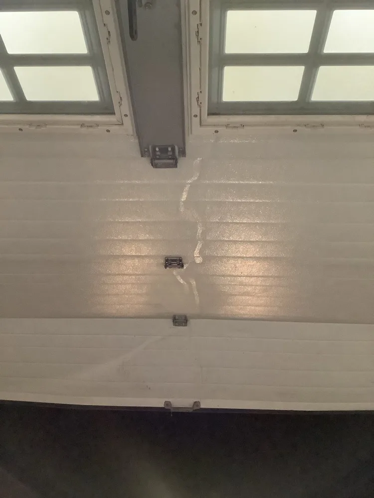 The upper sections of a white garage door seen from inside, a pale crease running down through two panels below the row of window lites where the door has been buckled.