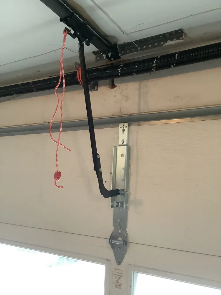 A garage door opener arm bolted through a reinforcing bracket on the top section of the door, the red and white emergency release cord hanging beside it and the torsion shaft running along the header behind.