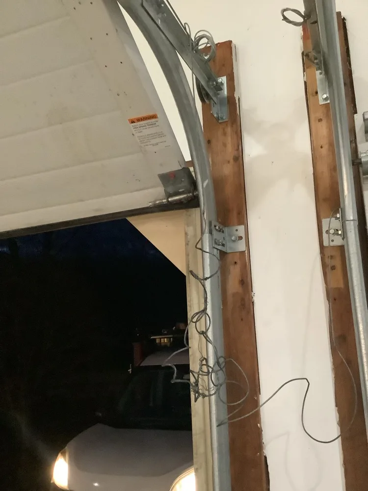 A garage door raised onto its curved track at night, with lift cable tangled in loose loops against the timber jamb and a car with its headlights on standing in the drive outside.