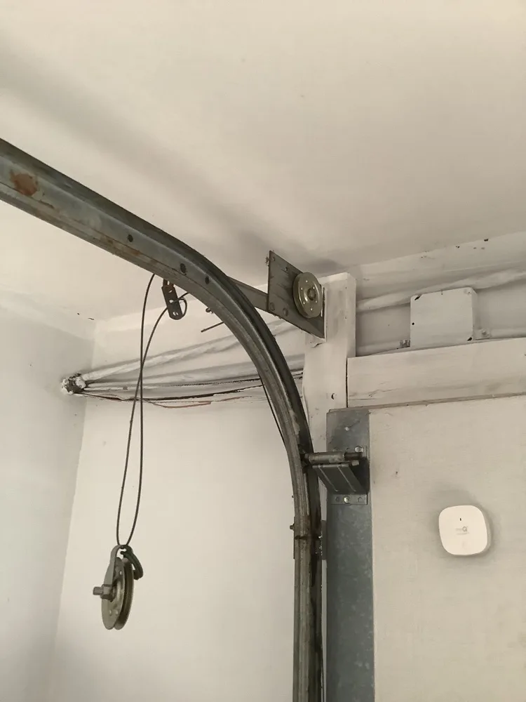 The curved section of a garage door track with its cable bracket at the ceiling, a pulley hanging loose halfway down on slack lift cable instead of sitting under tension.