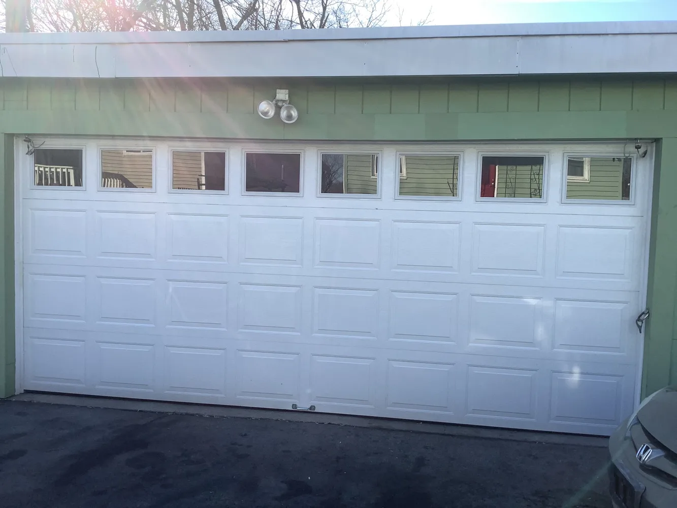 A white raised-panel double garage door with a band of eight window lites, set in pale green siding under a low winter sun, bare branches and clear sky above the roofline.