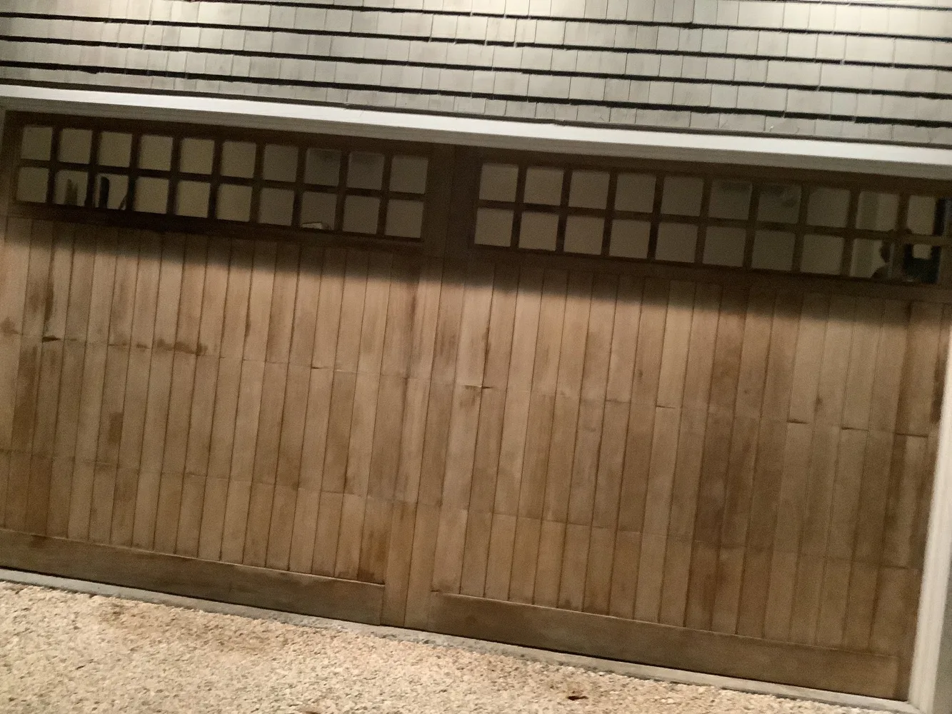 A stained wood carriage-style garage door with two rows of divided window lites, set beneath weathered cedar shingle siding and a white trim board.